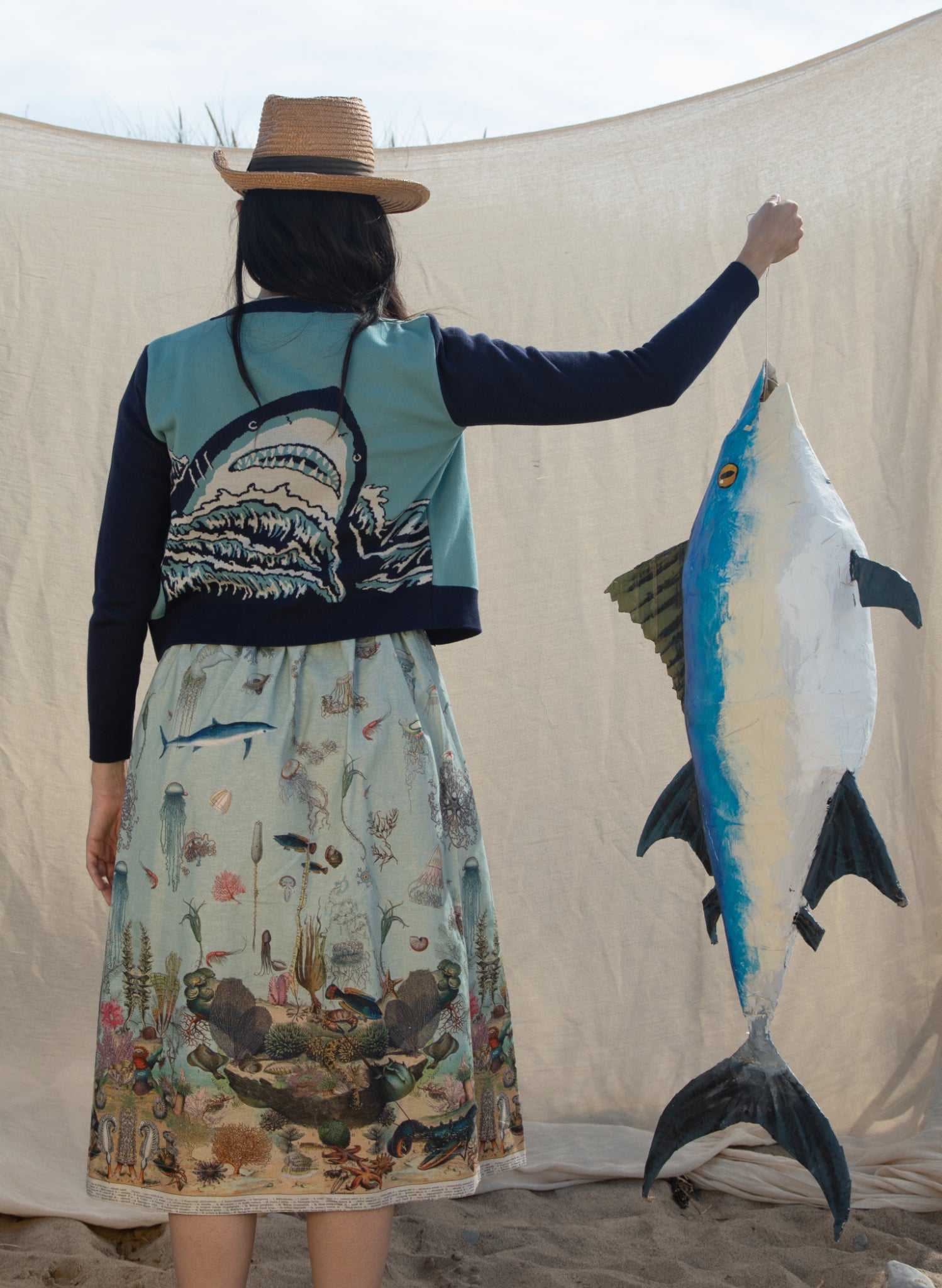 picture of a woman's back, wearing a cardigan featuring a shark head looming over some waves. The woman is wearing a summer hat and she is holding a large papier mache tuna fish.