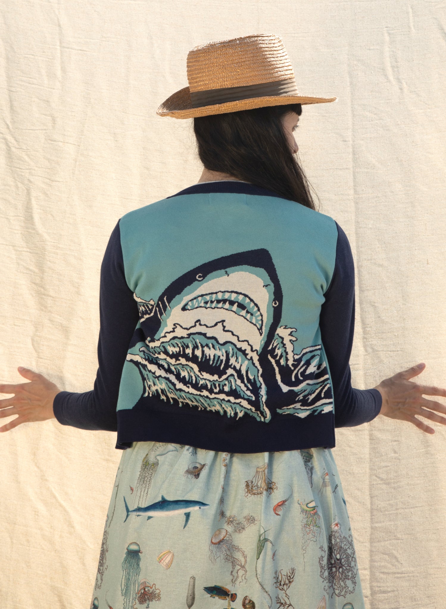 A picture of a woman from behind. She is wearing a cardigan featuring a sharks head looming over some waves. She wears a long skirt featuring vintage illustrations of aquatic life. The woman is wearing a summer hat looking to her right with her hands in front and palms splayed outwards touching a cream fabric backdrop lightly illuminated by the sun.