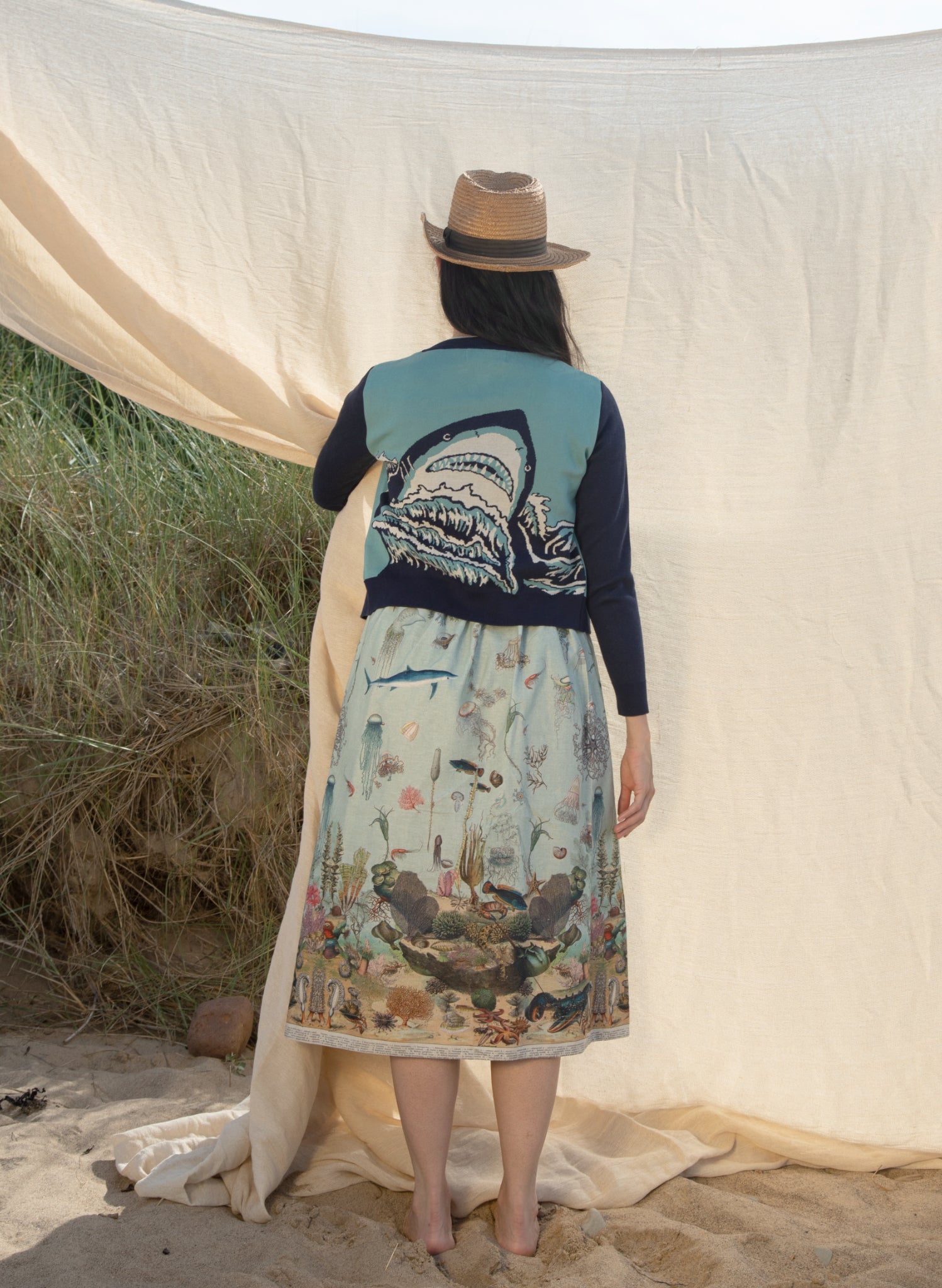 A picture of a woman from behind. She is wearing a cardigan featuring a sharks head looming over some waves. She wears a long skirt featuring vintage illustrations of aquatic life. The woman is wearing a summer hat and is standing in sand whilst holding backdrop fabric.